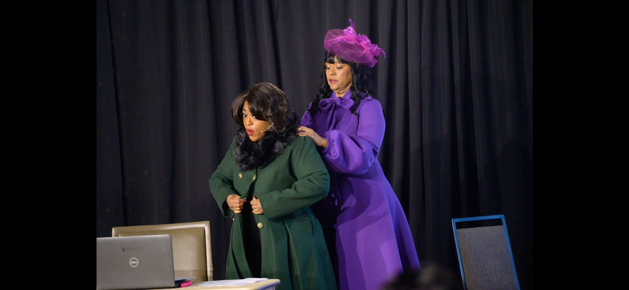 Two performers on stage in green and violet coats, a lifted-hat moment